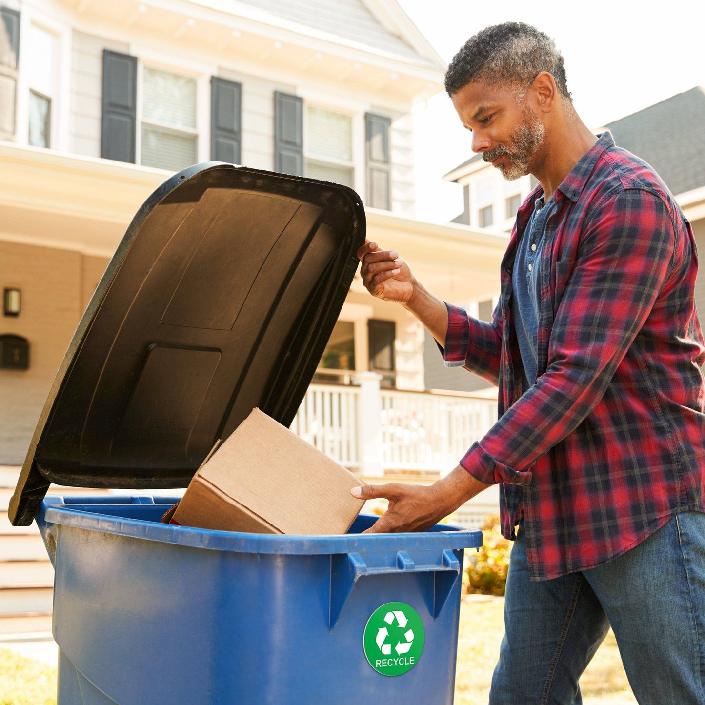 A man placing a cardboard box into a recycling bin with a green recycle sticker prominently displayed, illustrating the sticker's use on outdoor bins