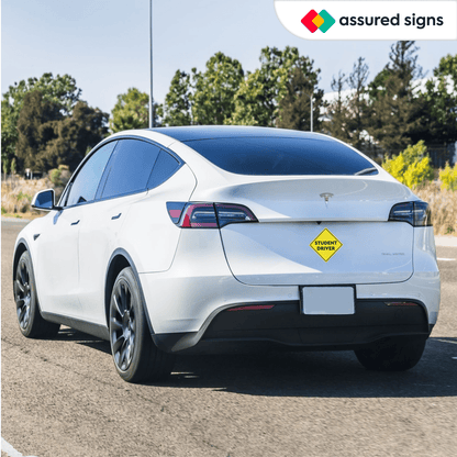 A yellow student driver car magnet placed on the rear of a white Tesla, promoting safety for new drivers on the road.